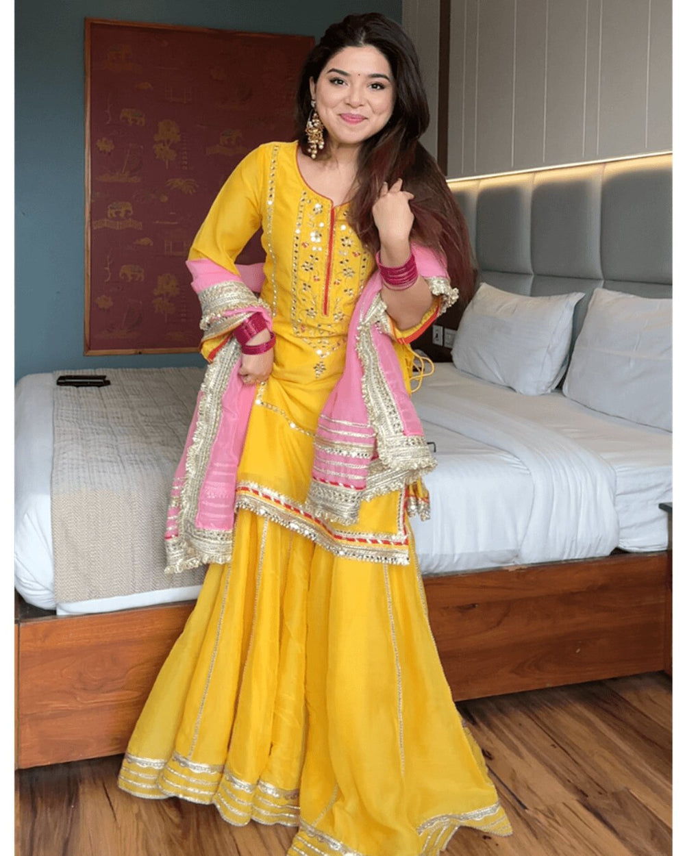 A woman models a traditional Indian salwar kameez outfit in bright yellow with pink sleeves and a pink dupatta, decorated with gold embroidery and mirror work, set against a bedroom background.