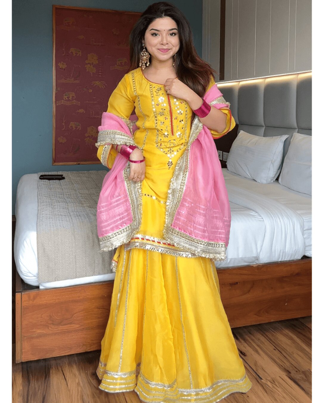 A woman models a traditional Indian outfit consisting of a bright yellow kurta and flared skirt with silver embroidery, paired with a pink dupatta featuring gold borders, in a bedroom setting.