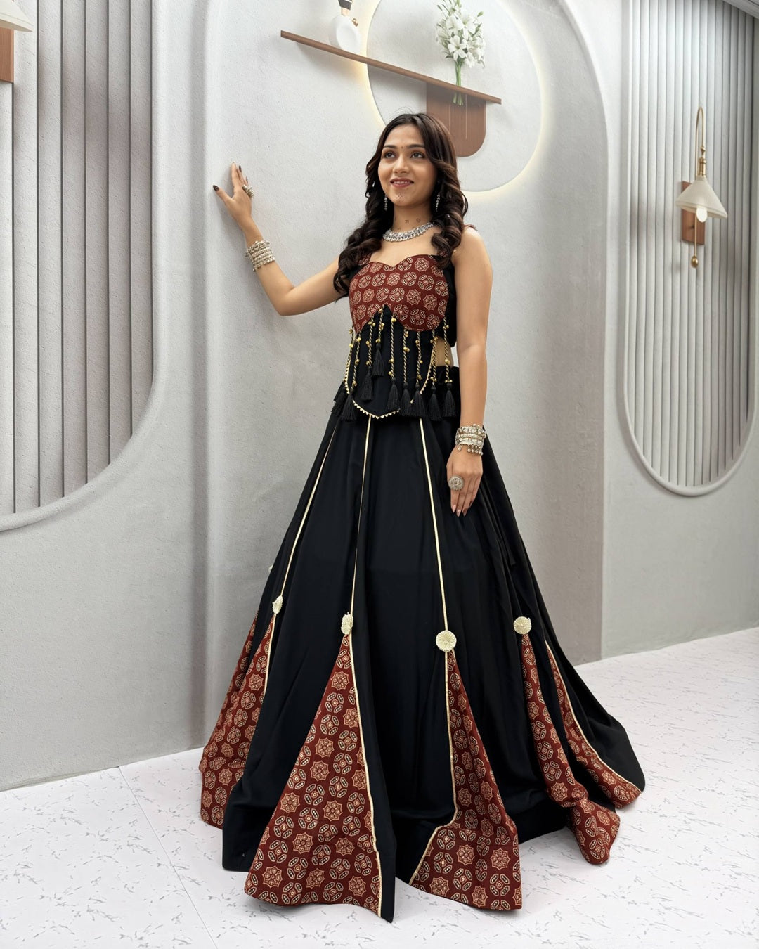 A woman models a traditional Indian evening gown with a black body, a brown patterned bodice and flared underskirt panels, and gold decorative accents, set against a modern gray architectural background.