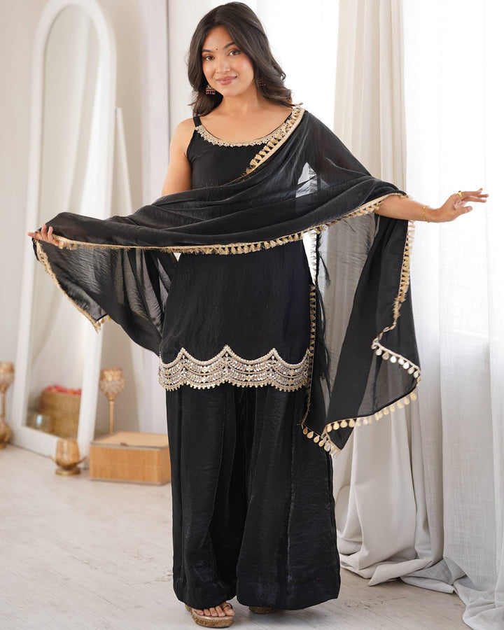 A woman modeling a black Indian ethnic suit set with gold embroidery, featuring a sleeveless top, wide-leg pants, and a matching dupatta, posed in a bright, modern room with white drapes.