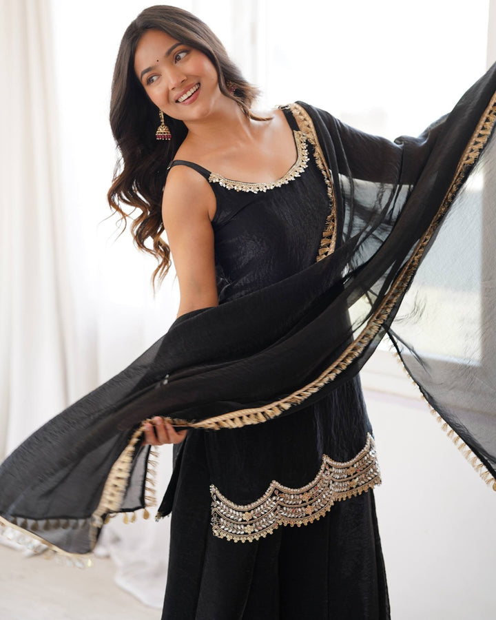A woman modeling a black traditional outfit with gold and silver embroidery, featuring a sleeveless top, wide-leg pants, and a matching sheer dupatta with gold trim.