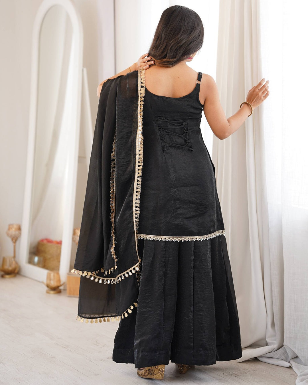 A woman shown from behind wearing a black traditional outfit with a matching dupatta draped over her shoulder, featuring gold embroidered borders and tassels, in a bright indoor setting.