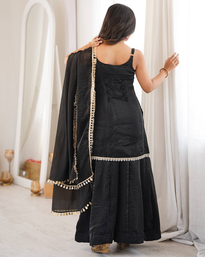 A woman shown from behind wearing a black traditional outfit with a matching dupatta draped over her shoulder, featuring gold embroidered borders and tassels, in a bright indoor setting.