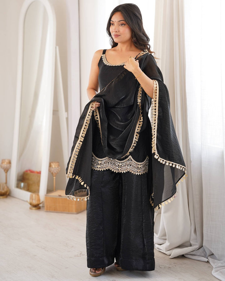 A woman models a black three-piece traditional outfit with wide-leg trousers, a sleeveless top, and a matching dupatta draped over her shoulder. The fabric features elegant gold and silver embroidery along the edges and neckline, set against a bright, softly lit indoor background with white curtains.