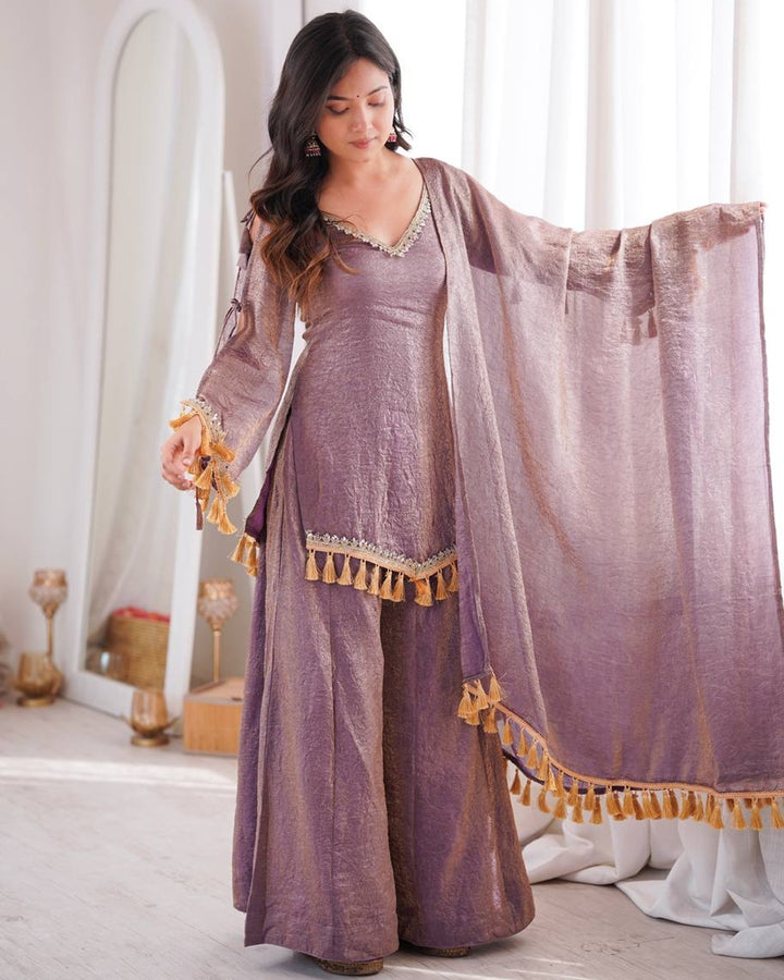 A woman models a traditional three-piece salwar kameez suit in a dusty purple/lavender color with gold tassel fringe and embroidery, paired with a matching sheer dupatta.