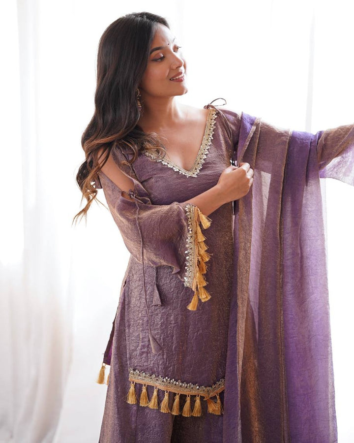 A woman wearing a purple traditional outfit with gold tassel trim and a matching sheer dupatta, standing in front of a bright white curtain.