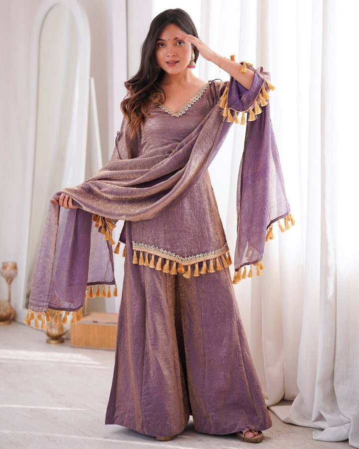 A woman models a lavender purple traditional outfit with wide-leg pants, a matching draped scarf, and mustard yellow tassels, set against a bright, softly lit indoor background with white curtains.