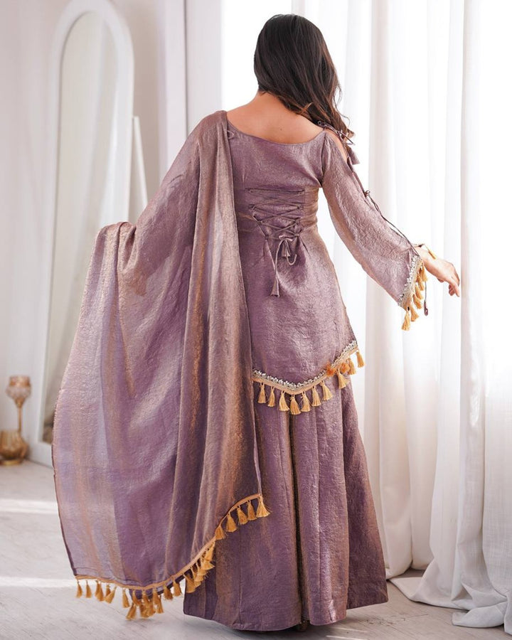 A woman wearing a lavender-purple traditional outfit with a matching sheer dupatta and gold tassels, photographed from behind in a bright indoor setting with white curtains.