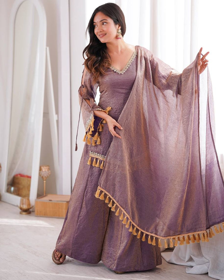 A woman models a traditional Indian outfit consisting of a dusty purple kurta, matching wide-leg pants, and a sheer purple dupatta with gold tassel trim, set against a bright, white-walled indoor background.