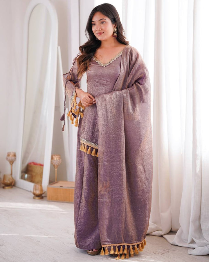 A woman models a traditional Indian outfit consisting of a lavender-purple kurta with gold embroidery and a matching draped dupatta with tassels, set against a soft, neutral indoor background with white curtains.