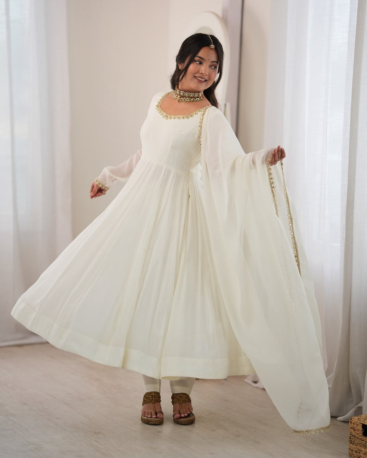 A woman modeling a traditional white Anarkali gown with gold embroidered details, a matching dupatta, and a white headpiece in a bright indoor setting with sheer curtains.