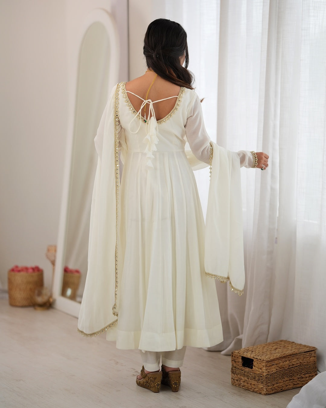 A woman viewed from behind wearing a cream-colored traditional dress with a draped dupatta, gold trim, and tie-back details, standing in a bright room with sheer curtains and a mirror.