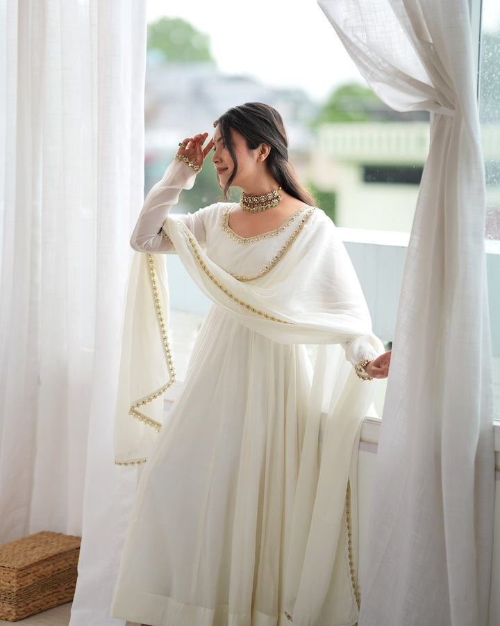 A woman wearing a white traditional outfit with gold trim and a matching dupatta, standing by a window with sheer white curtains.
