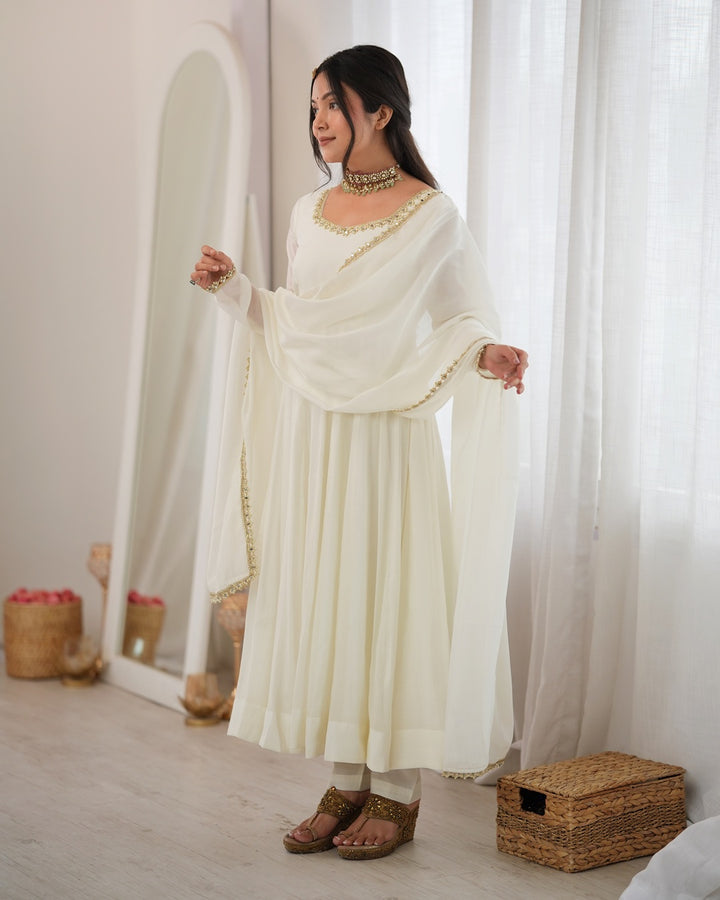A woman modeling a cream-colored traditional outfit with a matching dupatta, featuring gold embroidery details, standing in a bright, minimalist room with sheer curtains and a large floor mirror.