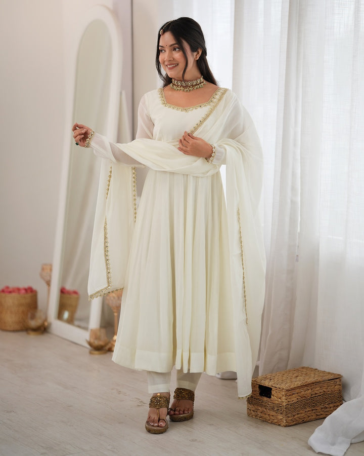 A woman modeling a traditional Indian outfit consisting of an off-white/cream kurta with gold embroidered borders, a matching dupatta, and white pants, standing in a bright room with sheer white curtains.