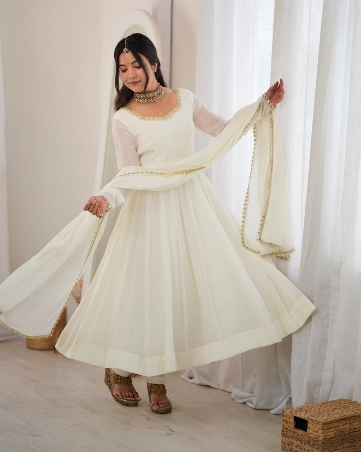 A woman modeling a traditional white Anarkali gown with sheer sleeves, gold embroidered details, and a matching white dupatta with gold trim, set against a soft, neutral indoor background with white curtains.