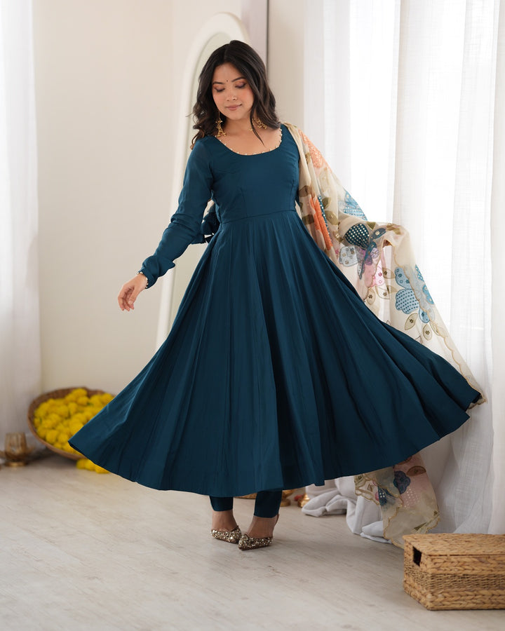 A woman modeling a long-sleeve, scoop-neck dark teal Anarkali-style dress with a flared skirt, paired with a patterned cream dupatta, in a bright, neutral indoor setting.