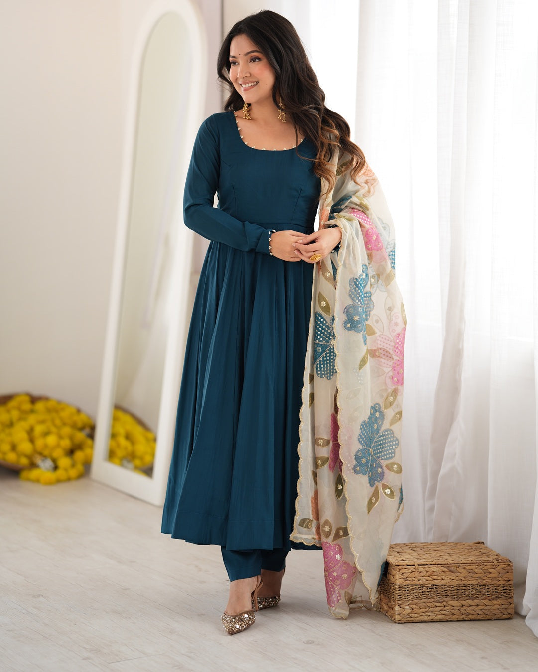 A woman models a dark teal blue long-sleeve kurta set with matching pants and a cream dupatta featuring colorful floral patterns, standing in a bright, modern room.