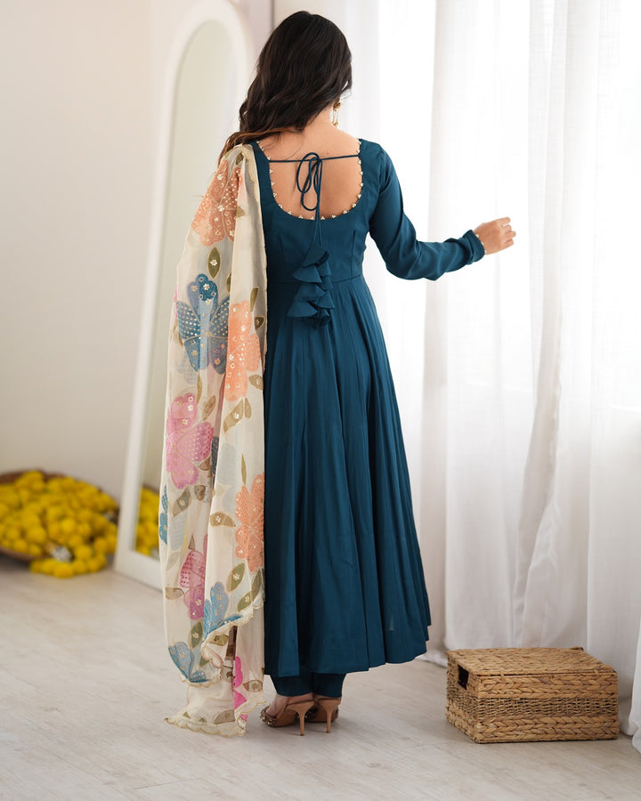 A woman stands with her back to the camera wearing a long-sleeved teal blue dress with a scalloped open back and a floral printed dupatta draped over her shoulder, in a bright room with white curtains.