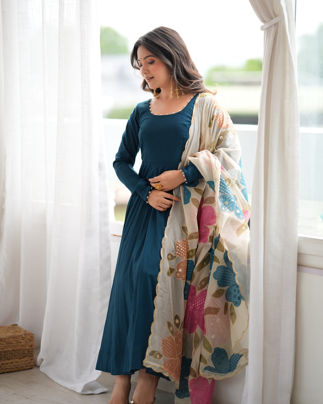 A woman models a teal blue long-sleeve kurta with a matching solid teal dupatta, while wearing a large cream dupatta with a colorful floral print. She is standing in front of a window with white sheer curtains.