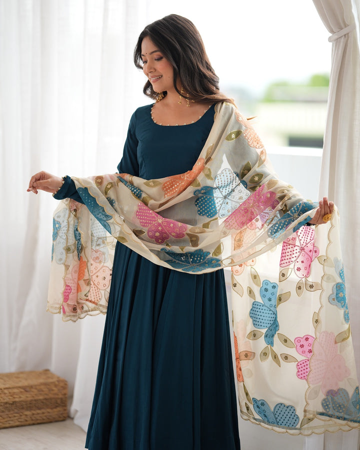 A woman wearing a dark teal traditional outfit with a long skirt and a matching sheer dupatta featuring colorful floral patterns in pink, blue, and beige, standing in front of a window with white curtains.