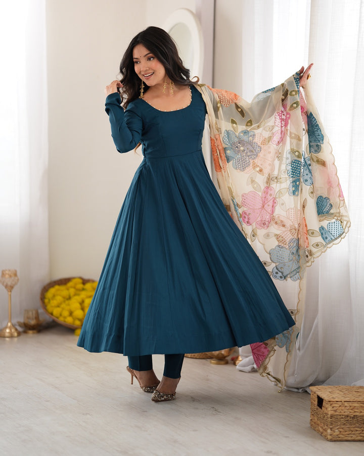 A woman models a long-sleeved teal blue maxi dress with a flared skirt, paired with a sheer floral dupatta, in a bright, modern room with white curtains.