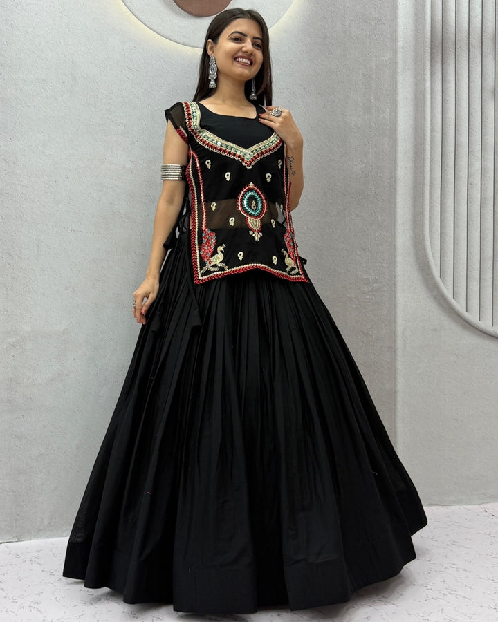 A woman modeling a black traditional Anarkali-style dress with a flared skirt and a heavily embroidered bodice featuring red, gold, and green details, set against a modern light gray wall.