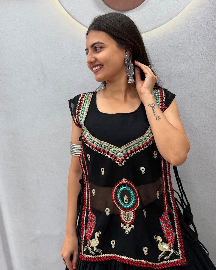 A woman wearing a black embroidered kurta with red, green, and gold beadwork and traditional motifs, standing against a light gray textured wall.
