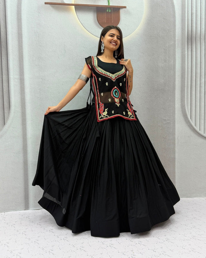 A woman modeling a black traditional dress with a heavily embroidered black bodice featuring red, gold, and green details, paired with a voluminous black flared skirt, set against a light gray textured studio wall.