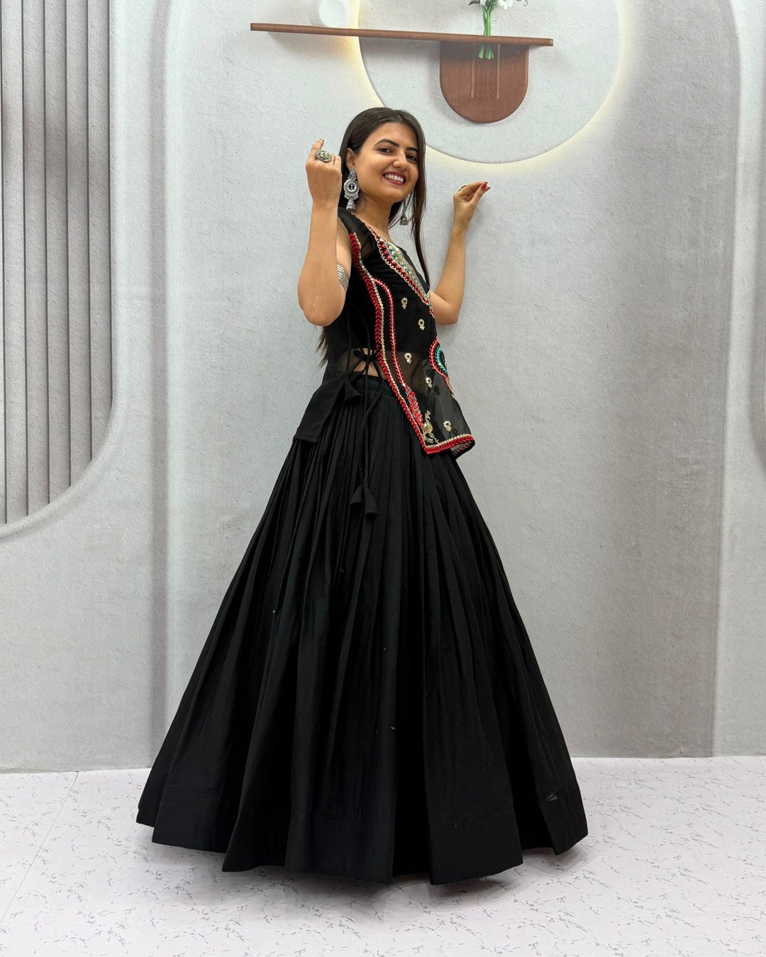 A woman modeling a black traditional Indian outfit consisting of a long flared skirt and a sleeveless embroidered vest, standing against a modern light gray wall.