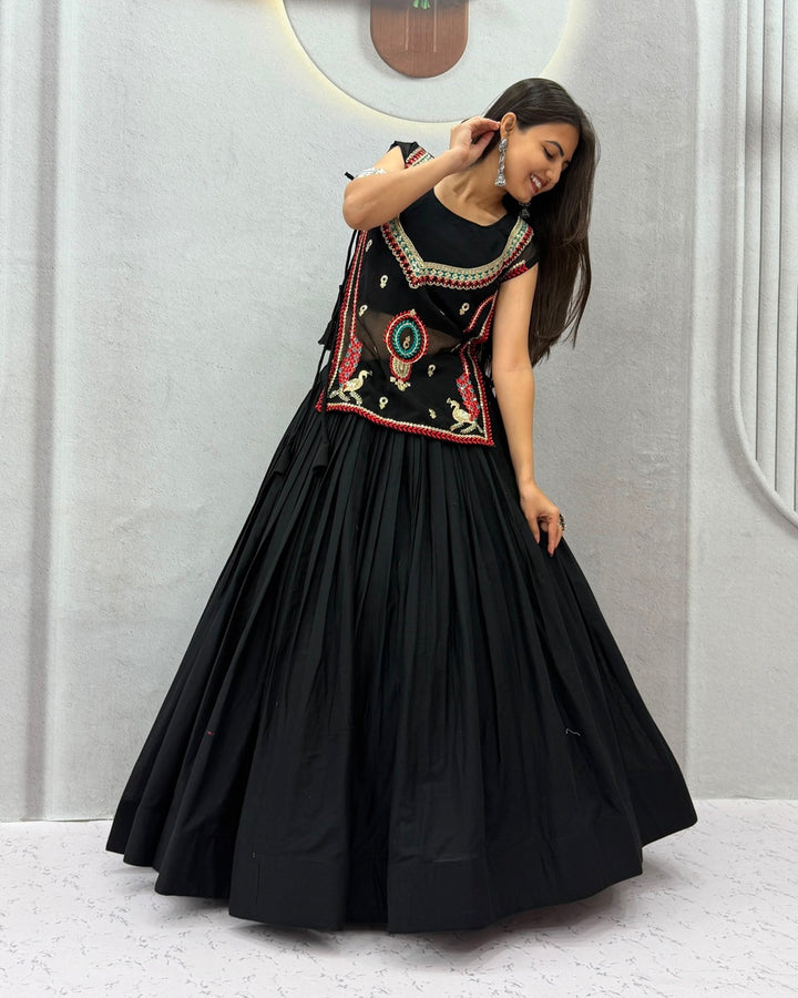A woman modeling a black traditional Anarkali-style dress with colorful embroidered details on the bodice and a full pleated skirt, standing against a light gray wall.