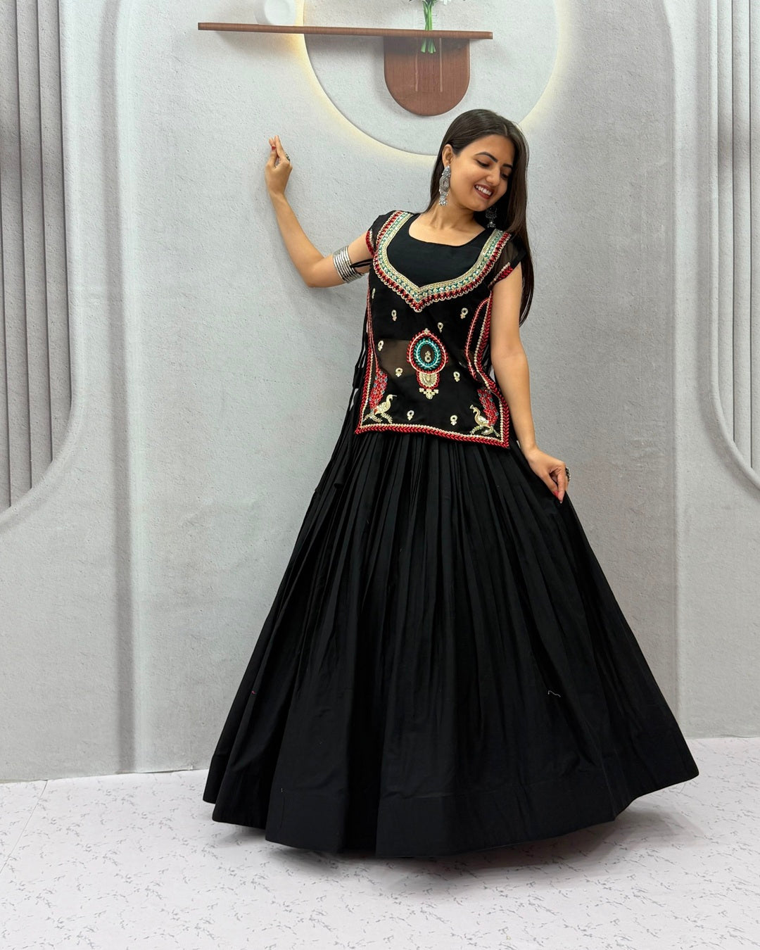 A woman modeling a black traditional long gown with a heavily embroidered bodice in red, gold, and green, paired with a full black flared skirt, set against a modern light gray wall with decorative arches.
