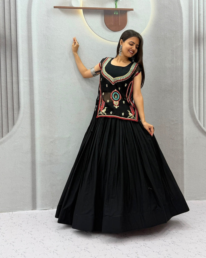 A woman modeling a black traditional long gown with a heavily embroidered bodice in red, gold, and green, paired with a full black flared skirt, set against a modern light gray wall with decorative arches.