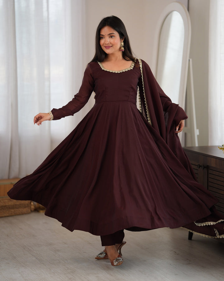 A woman modeling a long-sleeved maroon Anarkali gown with a matching dupatta in a bright indoor setting with white curtains.
