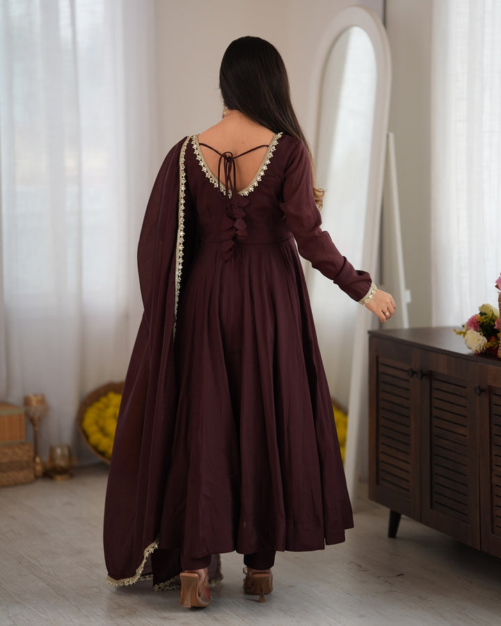 A woman wearing a long-sleeved maroon Anarkali gown with a draped dupatta, shown from the back in a bright, elegant room with white curtains and a wooden cabinet.