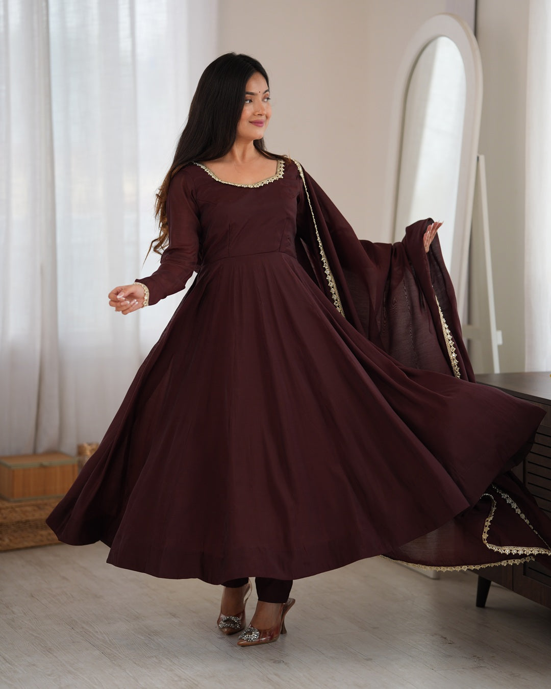 A woman wearing a long-sleeved maroon Anarkali gown with gold embroidery and a matching dupatta, captured in a bright indoor setting with a mirror in the background.