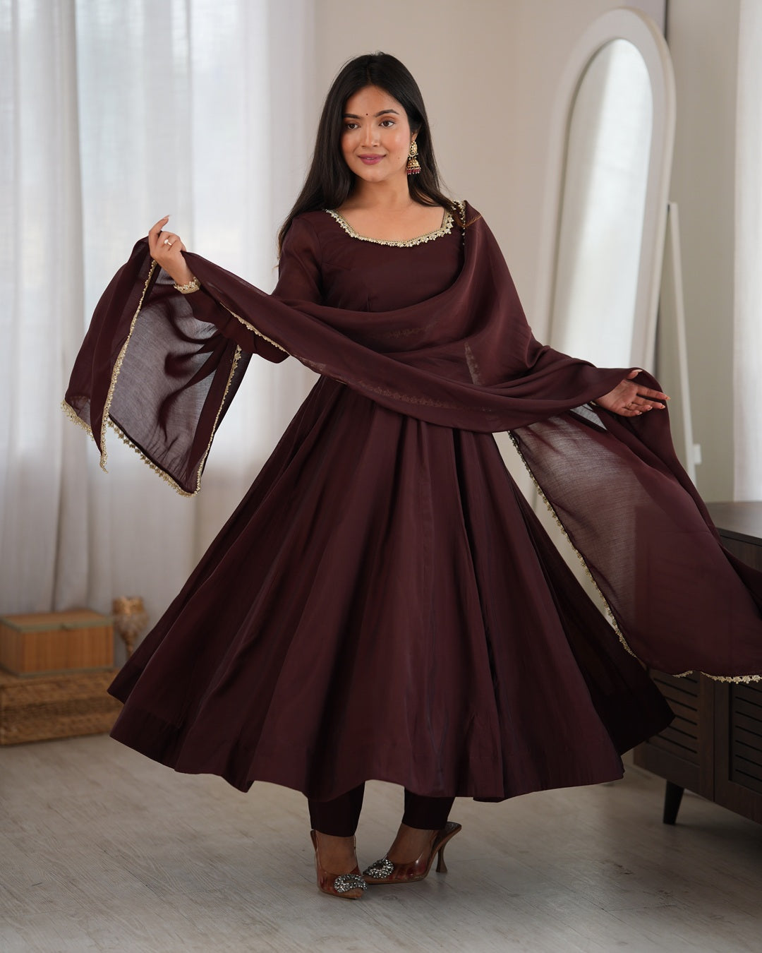 A woman modeling a traditional deep maroon Anarkali suit with a matching dupatta, featuring gold embroidered details on the neckline and dupatta borders, set in a bright indoor room.