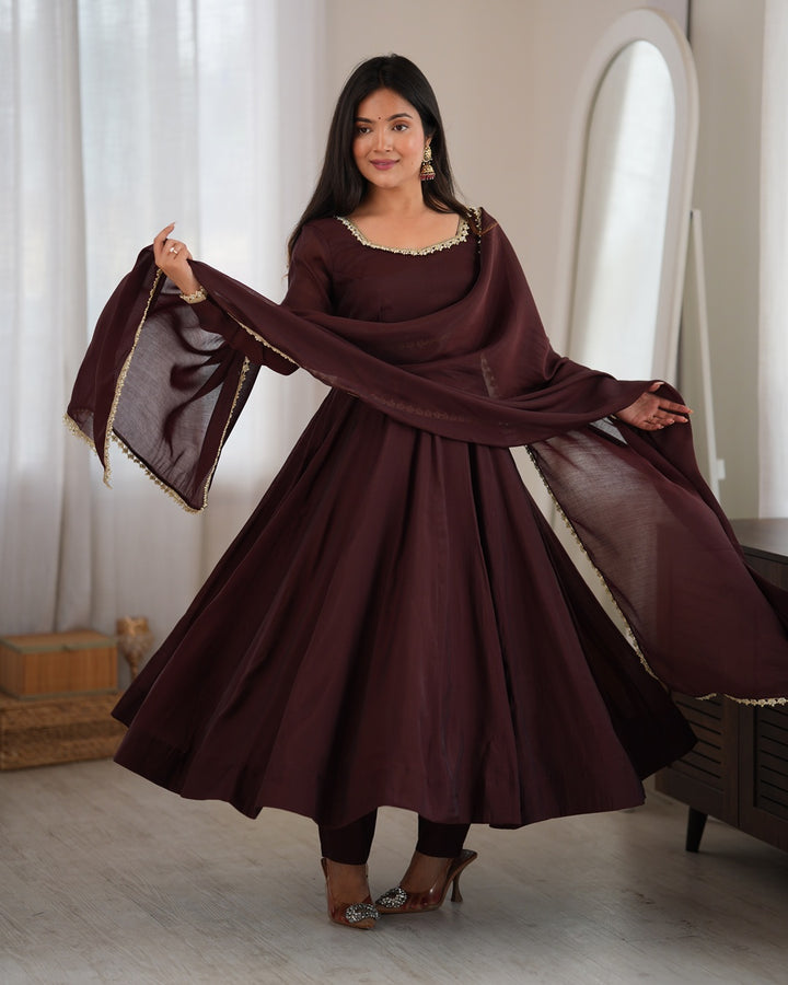 A woman modeling a traditional deep maroon Anarkali suit with a matching dupatta, featuring gold embroidered details on the neckline and dupatta borders, set in a bright indoor room.